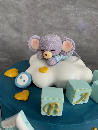 A blue cake topped with a sculpted sleeping teddy bear on a cloud, gold hearts, a button, and mint alphabet blocks lettered A and B.