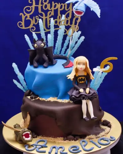 Two-tier cake with a 'Happy Birthday' topper, a black dragon figure, a girl in a Batman dress, a gold '6', and a name piped in blue.