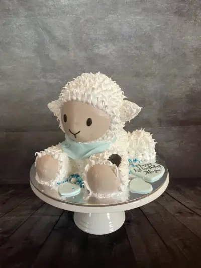 A white fluffy lamb-shaped cake with a pale blue scarf, sitting among light blue hearts, one with a first-birthday message.
