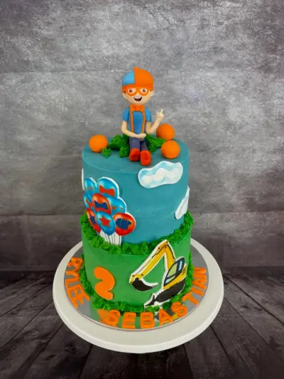 A two-tier cake with a boy figure in glasses and cap, balloons, a digger graphic, a number 2, and names piped round the base.