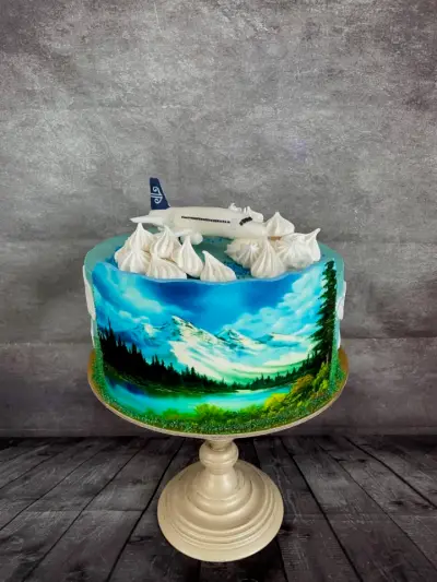 A cake with an airbrushed mountain and lake scene around the side, topped with a white model airplane and white meringue kisses.