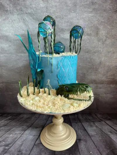 A teal cake with a sandy crumb base, edible shells, hanging glass-look orbs, a jagged fin decoration, and a sculpted green dinosaur skull.
