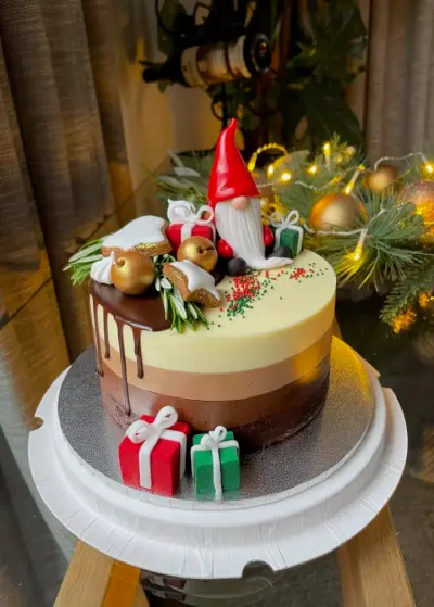 An ombre drip cake with a fondant gnome, gold baubles, mini pies, rosemary, and red and green gift boxes and sprinkles.