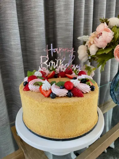 A crumb-coated cake topped with pink piped rosettes, strawberries, blueberries, raspberries, mint, and a 'Happy Birthday' topper.