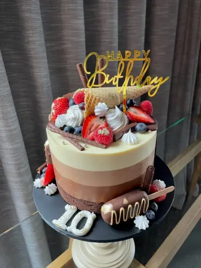 An ombre drip cake with berries, a waffle cone, chocolate sticks, a cakesicle, gold leaf, a number 10, and a 'Happy Birthday' topper.
