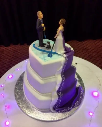 Three-tier white wedding cake with a heart-shaped top tier, a bride and groom paddleboarding topper with a cat, and a purple satin drape.