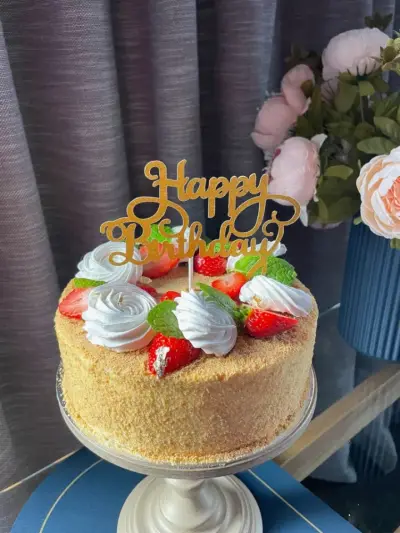 A golden crumb-coated cake topped with white piped rosettes, strawberries with gold leaf, mint, and a gold 'Happy Birthday' topper.