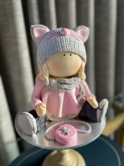 Sugar figurine of a girl in a pink dress with a grey knitted cat-ear hat, scarf, boots and a small pink purse.