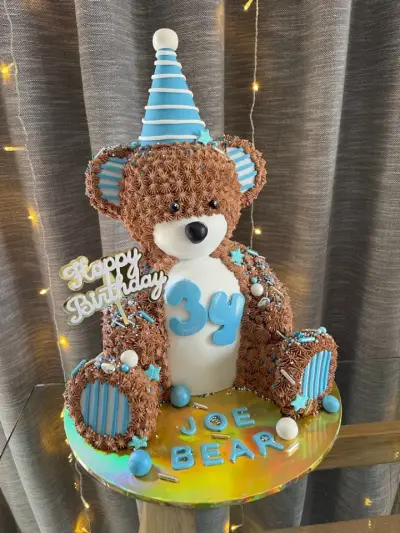 Bear-shaped cake with brown piped fur, a blue striped party hat and a '3y' number, with Joe Bear written on the base.