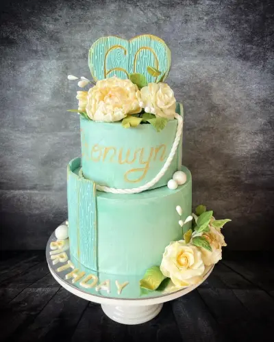 Two-tier mint cake with wood-grain panels, a gold '60' heart topper, cream flowers, a name in gold script and Birthday piped on the base.