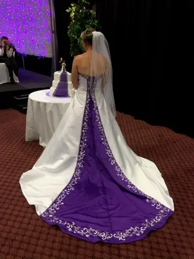 A three-tier white and purple wedding cake pictured at the reception, with the bride's purple-trimmed gown and veil in the foreground.