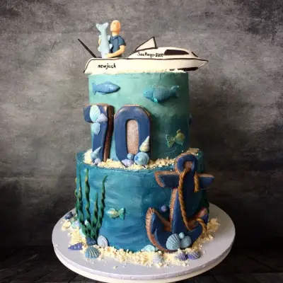 Two-tier ocean-themed cake with a '70', a boat and fisherman topper, fish, shells and an anchor decoration.