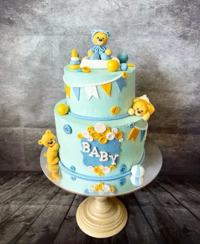 Two-tier mint green cake with teddy bear figures, bunting, buttons, and 'BABY' spelled on a blue gingham plaque.