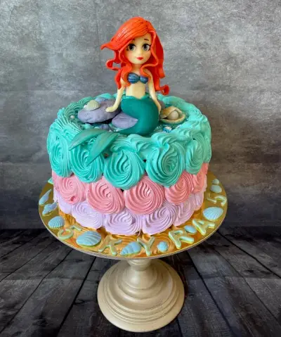 Round cake with turquoise, pink, and lavender rosette icing, a fondant mermaid on top, and shells with a number 5 around the base.