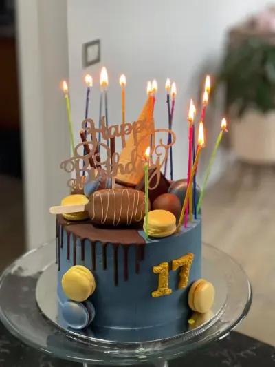 Navy cake with a chocolate drip, macarons and chocolate treats on top, a gold '17', and many lit rainbow-coloured candles.