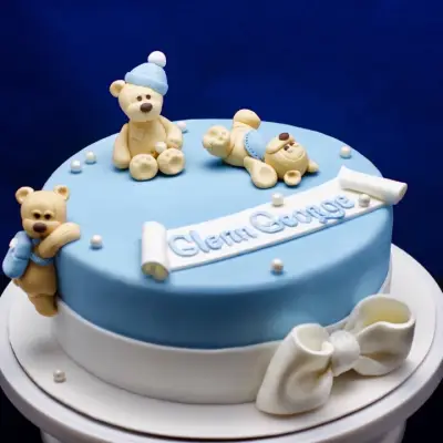 Pale blue round cake with a white base band, three teddy bear figures, a piped name scroll, pearl beads, and a white fondant bow.