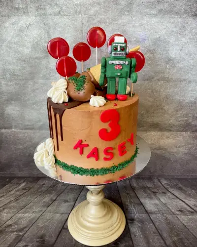 Caramel cake with a chocolate drip, green sprinkles, a red '3' and a name in fondant, plus a toy robot and red lollipops on top.