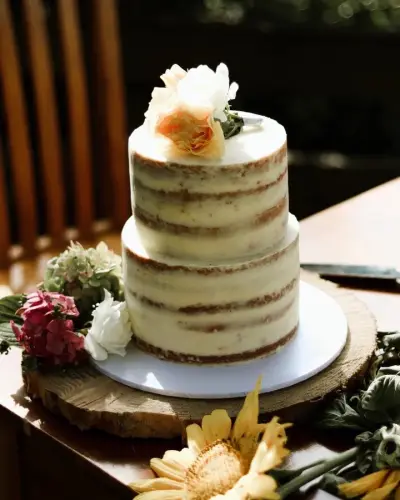 Two-tier semi-naked cake with thin buttercream, fresh peach and white flowers on top, displayed on a wood slice with a floral arrangement.