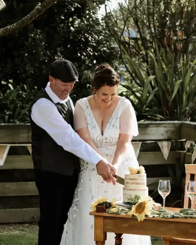 A bride and groom cut a semi-naked two-tier cake together outdoors, with sunflowers and bunting in the background.