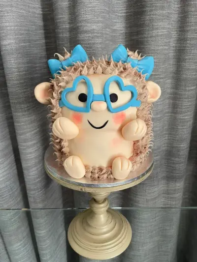 Cake shaped like a smiling hedgehog with heart-shaped blue glasses, blue bows, pink cheeks, and piped spike fur.