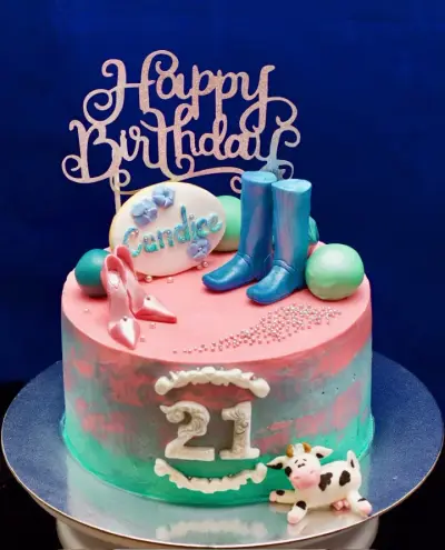 Pink and teal watercolour icing cake with a glitter 'Happy Birthday' topper, a name plaque, pink heels, blue boots, a cow, and '21'.