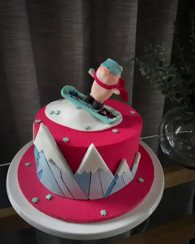 A hot pink cake with a snowy white top, fondant mountains around the base, and a fondant pig in teal goggles riding a snowboard.