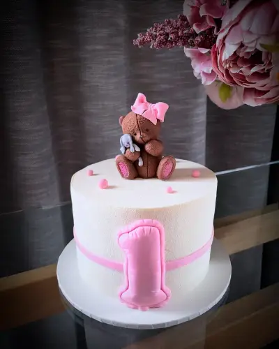White cake with a pink ribbon and a puffy pink number one, topped with a fondant teddy bear in a pink bow holding a toy bunny.