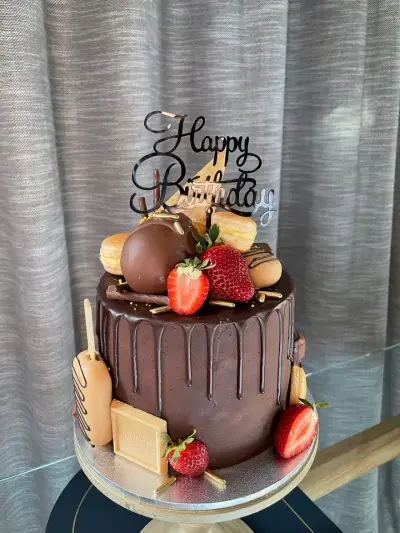 Dark chocolate drip cake topped with a waffle cone, chocolate truffles, macarons and strawberries beneath a Happy Birthday topper.
