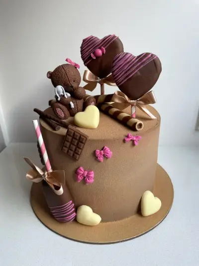Caramel cake decorated with a fondant teddy bear, two chocolate heart cake pops, chocolate hearts, wafer rolls and pink bows.