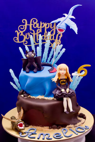 Two-tier blue and brown dragon-themed sixth birthday cake with an edible sculpted blonde girl in a black dress, black dragon, pale-blue flying dragon, smaller dragon, crystalline blue decorations and gold birthday topper.