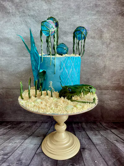 Tall turquoise Subnautica-themed cake with a sculpted green sea-creature head, edible sand and shells, underwater plants, translucent blue sugar fins and three hollow isomalt bubbles suspended within trailing green seaweed.